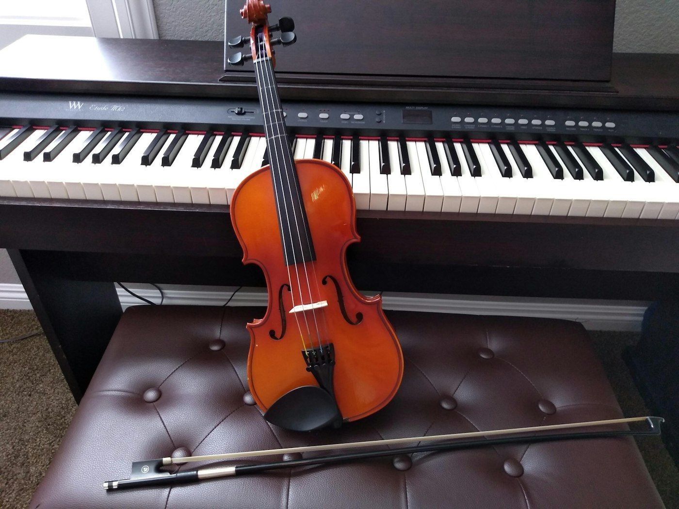 Violin and bow on a padded bench in front of a piano. The violin is brown.