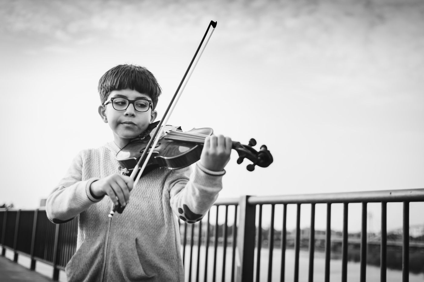 Boy playing a violin outdoors, wearing glasses, holding the bow, against a cloudy sky and metal railing.