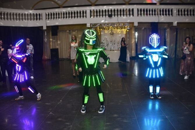 Tres bailarines con trajes de neón y luces LED brillantes actúan en un escenario interior oscuro.