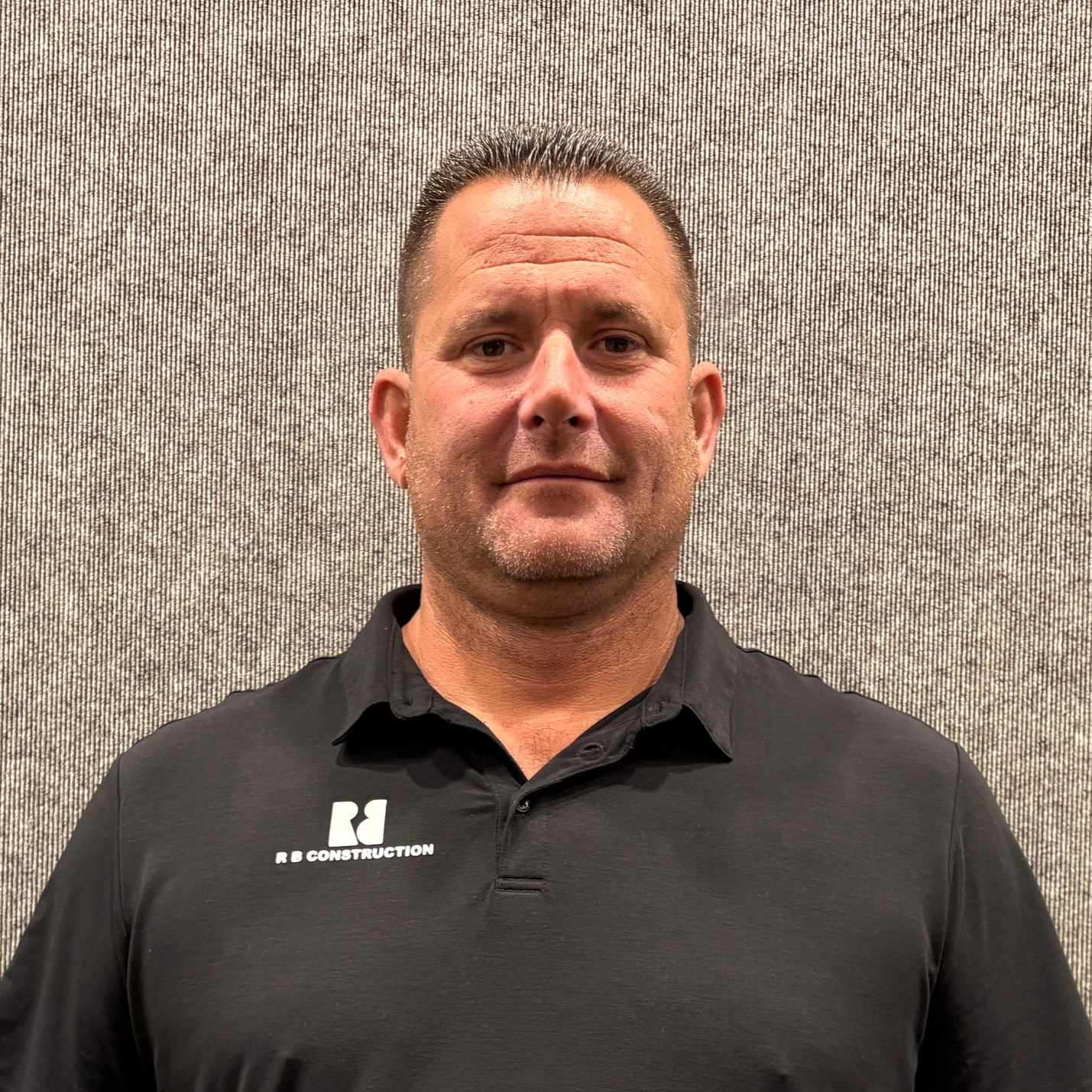 Man in a black polo shirt standing against a textured gray wall, facing forward with a slight smile.