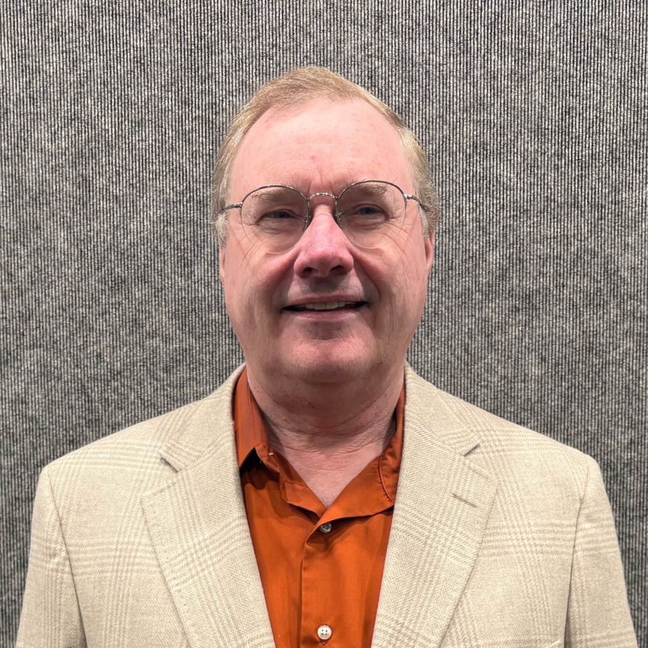 Man in beige blazer and orange shirt standing against a gray textured wall, smiling at the camera