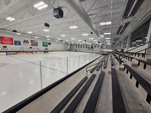 Empty indoor ice rink with white ice, black bleachers, and bright overhead lights