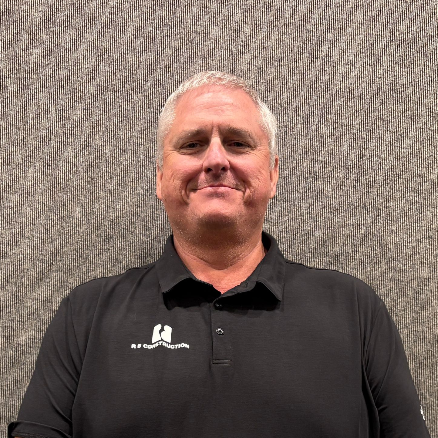 Smiling man in a black polo shirt standing against a textured gray wall