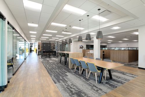 Modern office break area with long table, chairs, carpet, and glass walls under bright ceiling lights