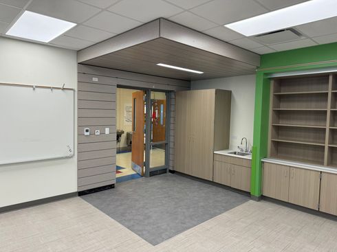 Empty classroom with whiteboard, gray carpet, wood cabinets, and a doorway to a hallway