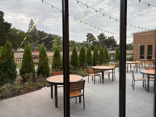 Outdoor patio with round wooden tables, benches, string lights, and tall evergreens beside a brick building