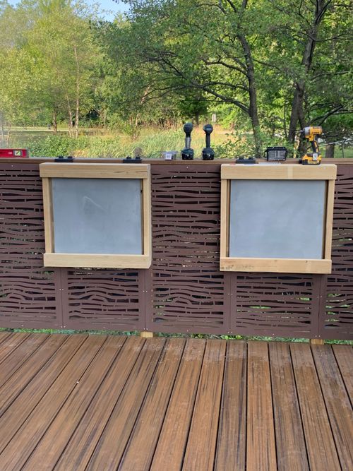 Outdoor wooden counter with two empty display frames on a deck, with trees and bottles behind it