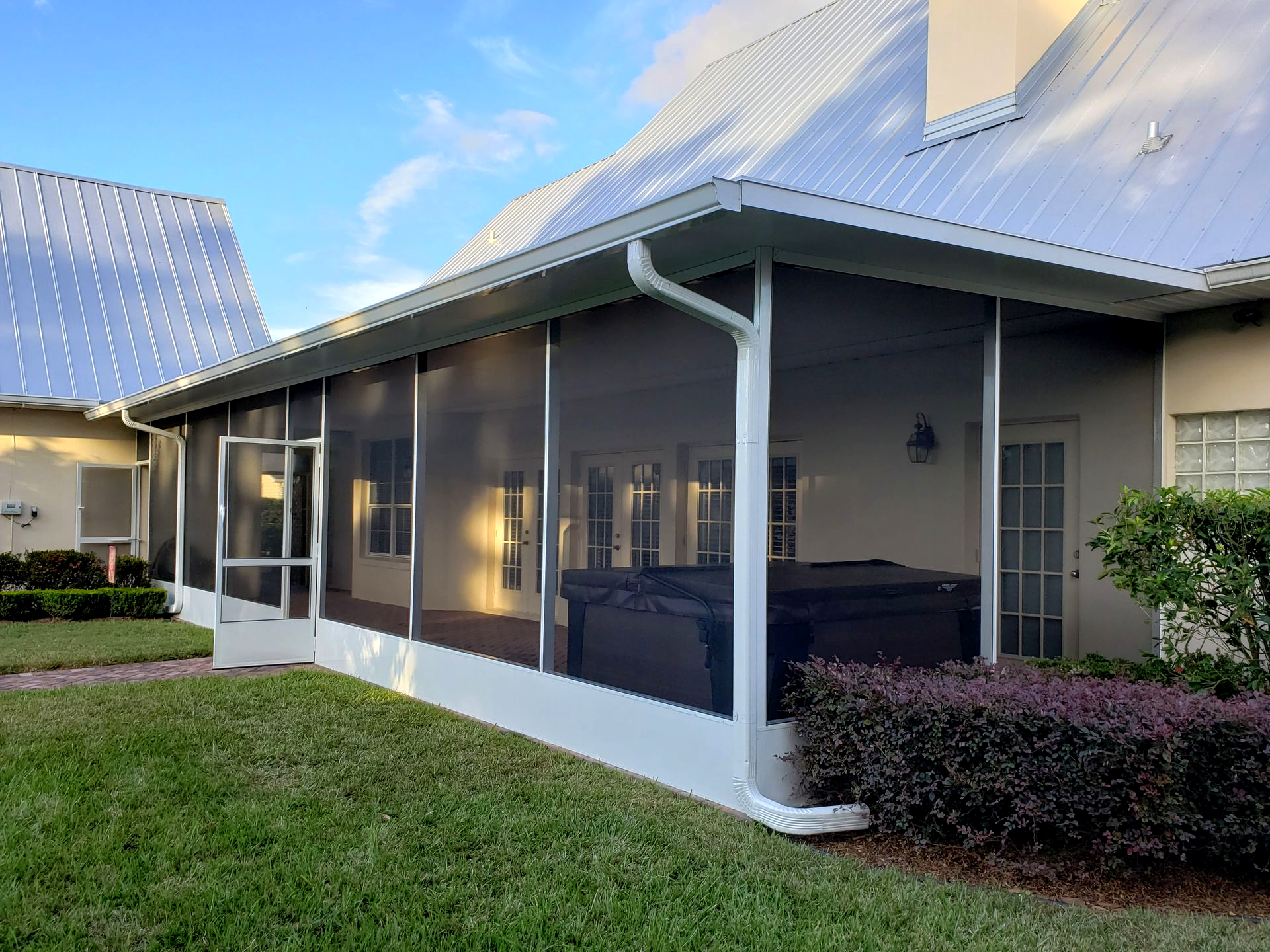 A white-framed screened-in porch attached to a house with a metal roof, featuring a hot tub on the patio.