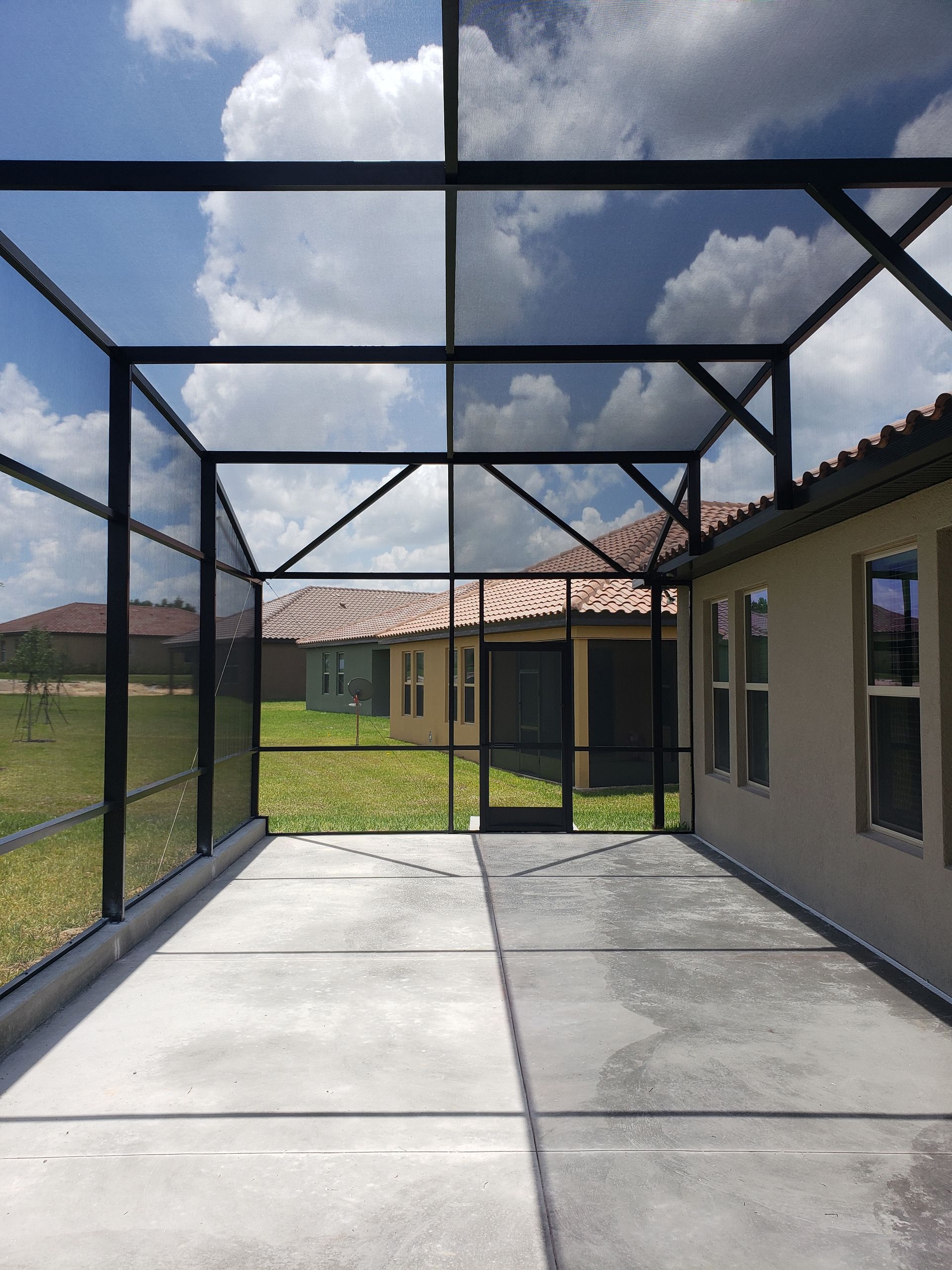 A screened-in patio with a concrete floor looking out towards a neighboring house under a partly cloudy sky.