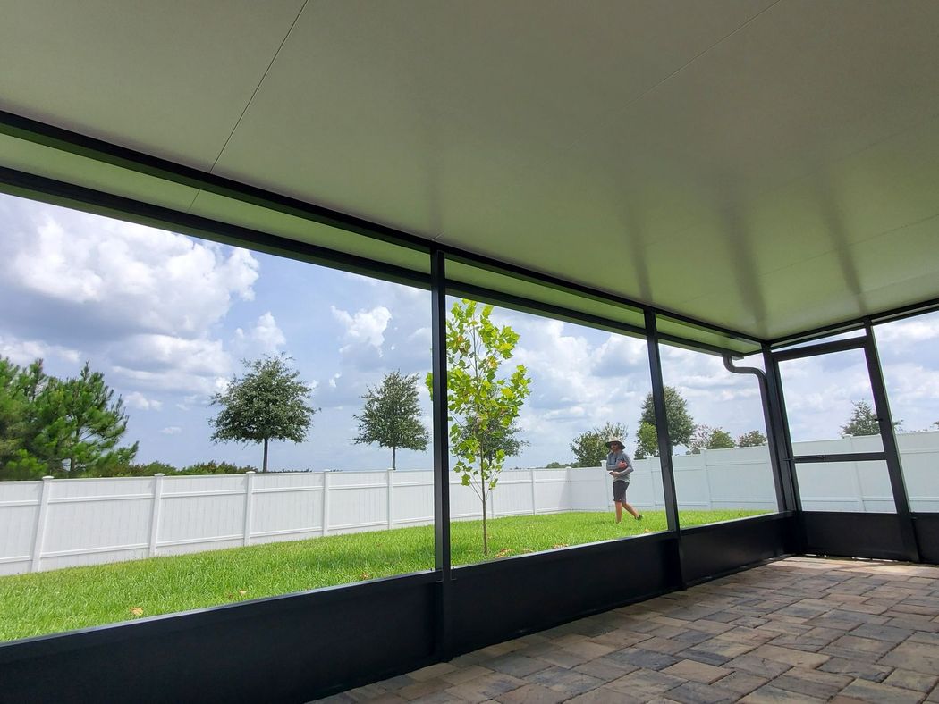 View from a screened-in patio overlooking a green lawn, white vinyl fence, and trees under a partly cloudy sky.