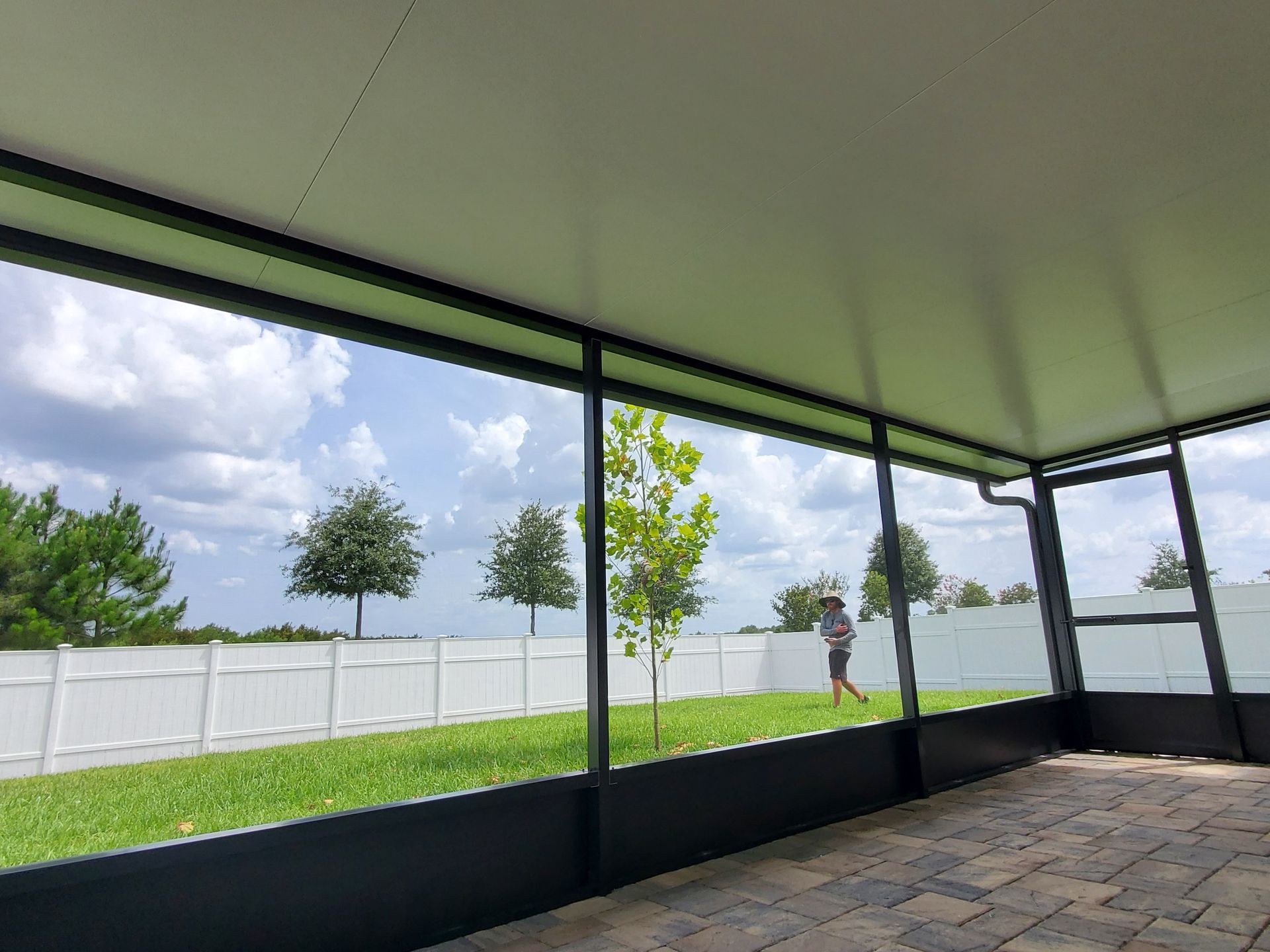 View from a screened-in patio overlooking a green lawn, white vinyl fence, and trees under a partly cloudy sky.