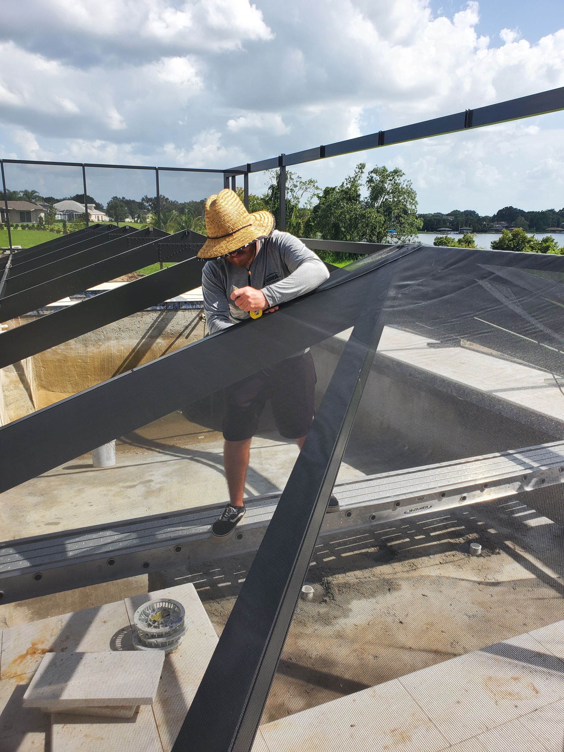 A person wearing a straw hat works on the aluminum frame and mesh screen of a residential pool enclosure.