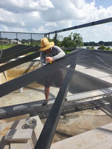 A person wearing a straw hat works on the aluminum frame and mesh screen of a residential pool enclosure.