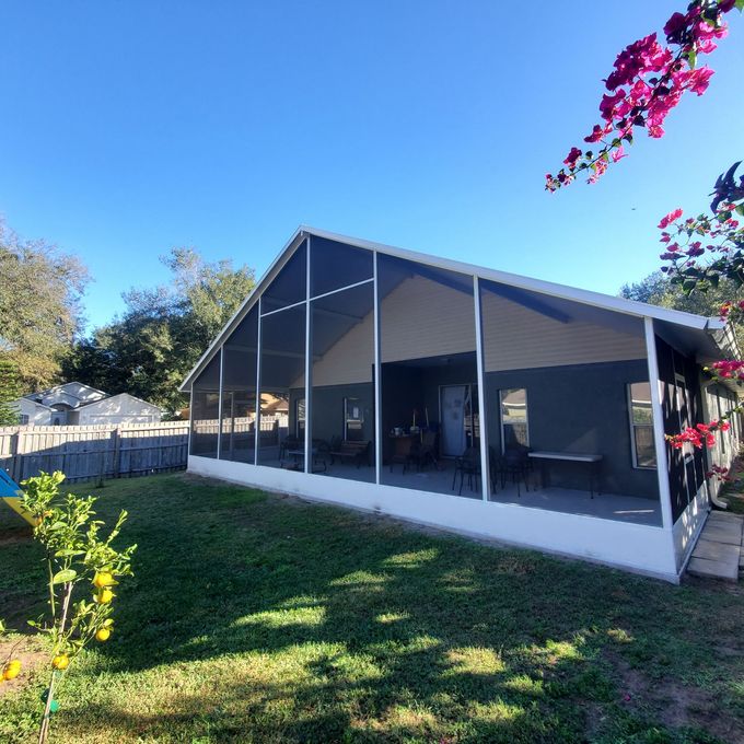 A screened-in porch with a gabled roof set in a grassy backyard, framed by tree branches and vibrant pink flowers.