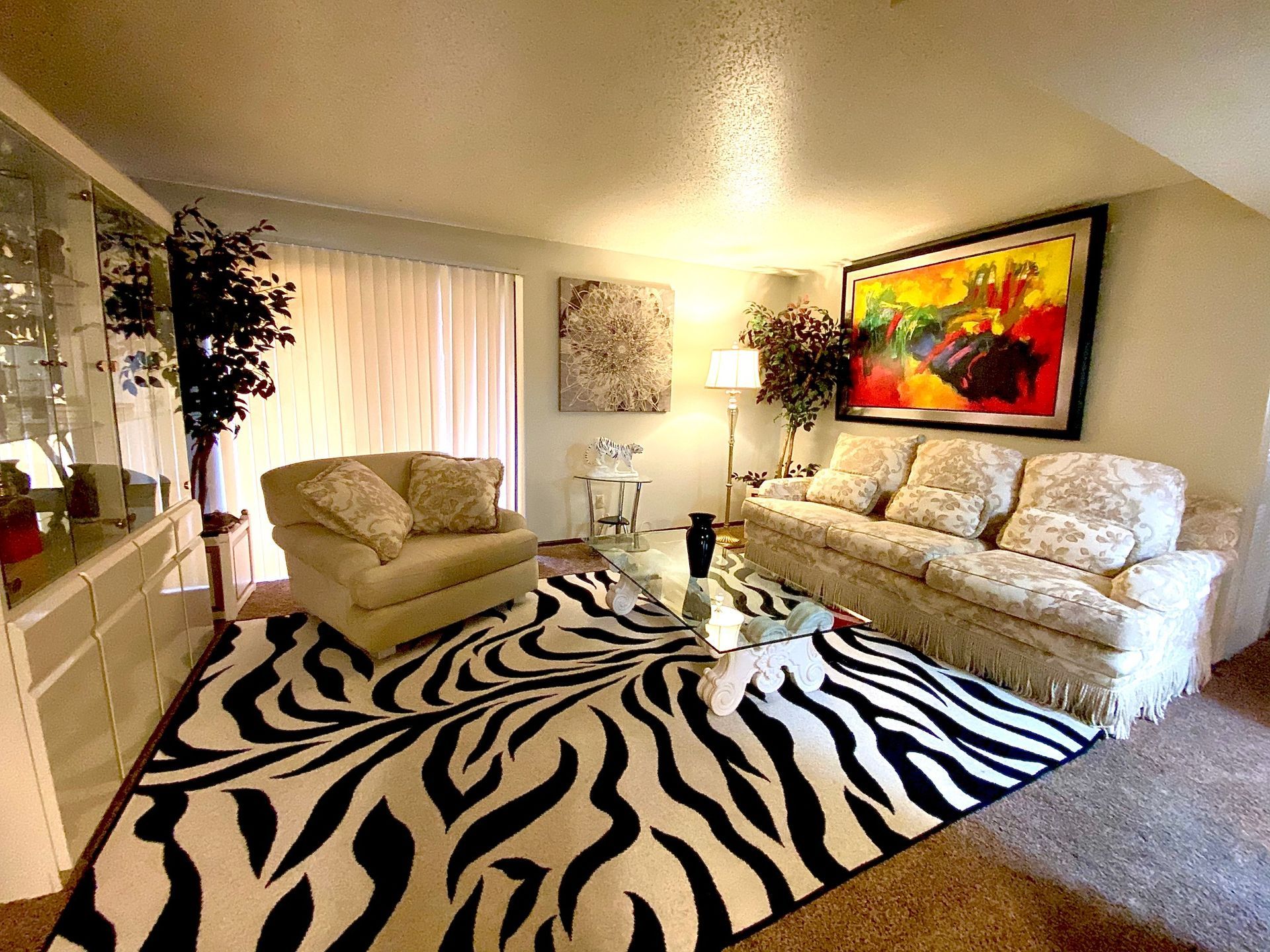 Living room with zebra-print rug, beige furniture, colorful abstract art, and large window with curtains.