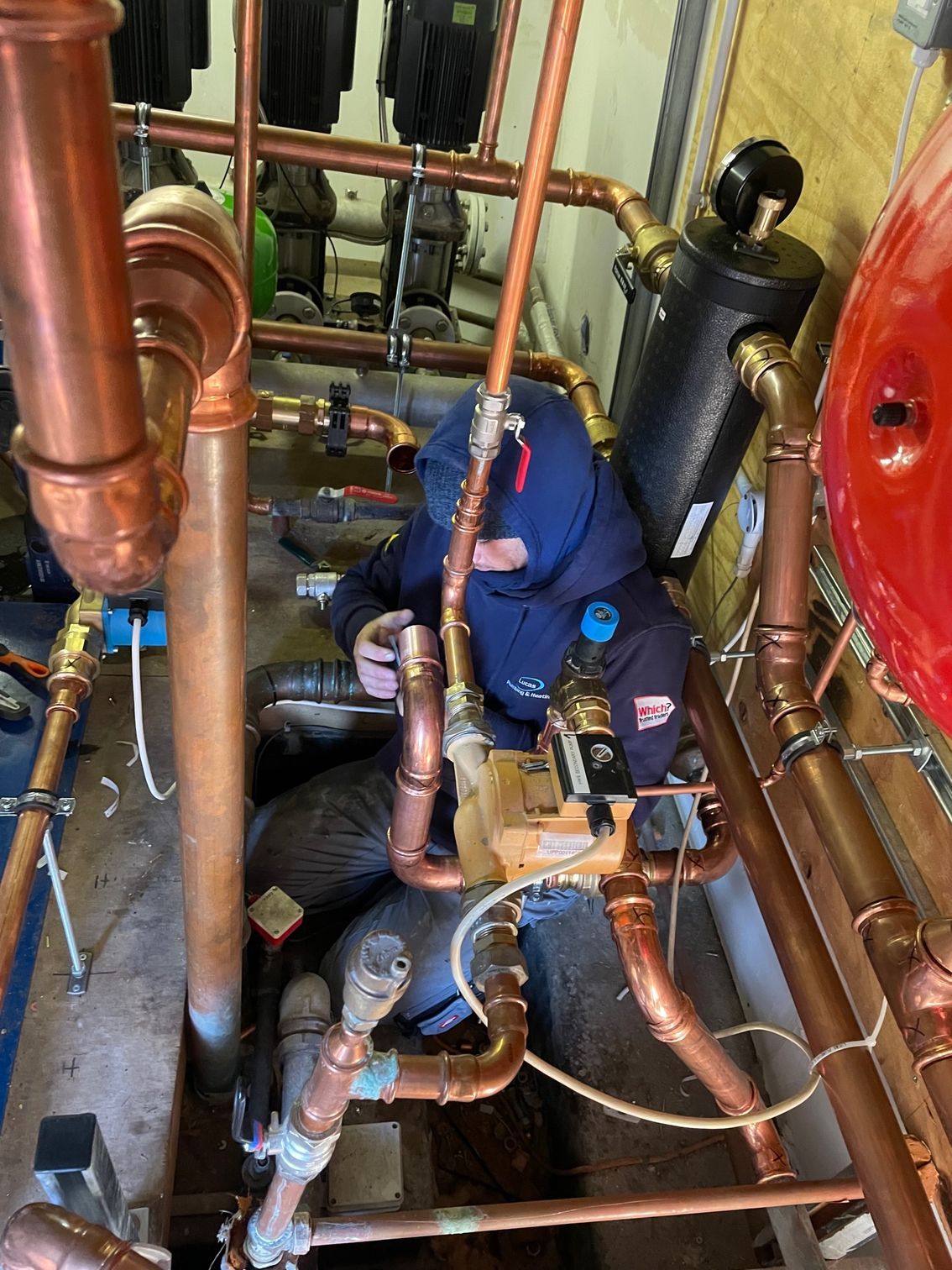 Plumber working on copper pipes in a tight space, wearing blue hooded jacket.