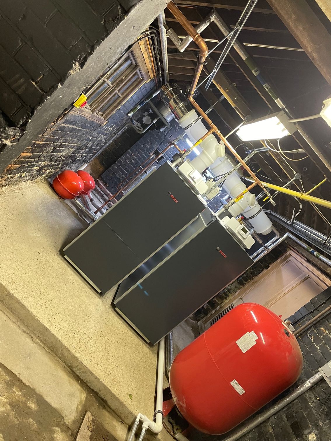 Two gray water heaters and two red tanks in a basement setting with pipes and exposed beams.