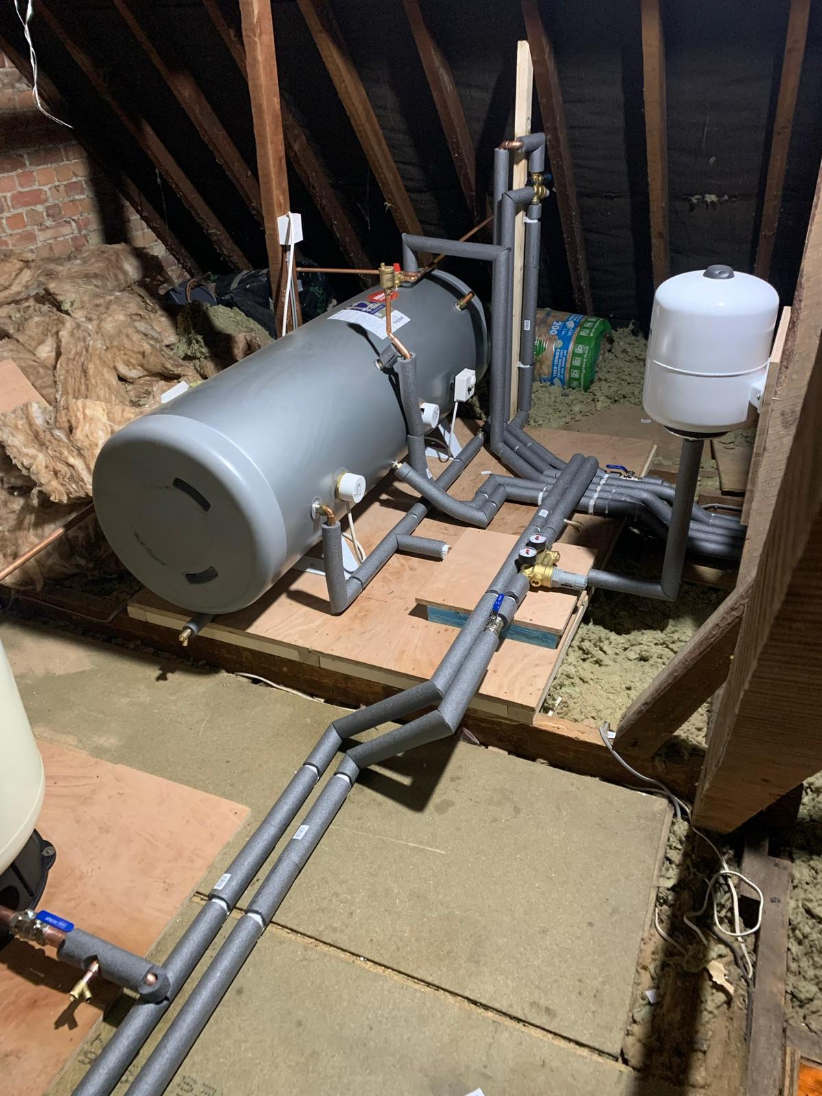 Large water tank and pipes in an attic, part of a heating system, with insulation and a white expansion tank.