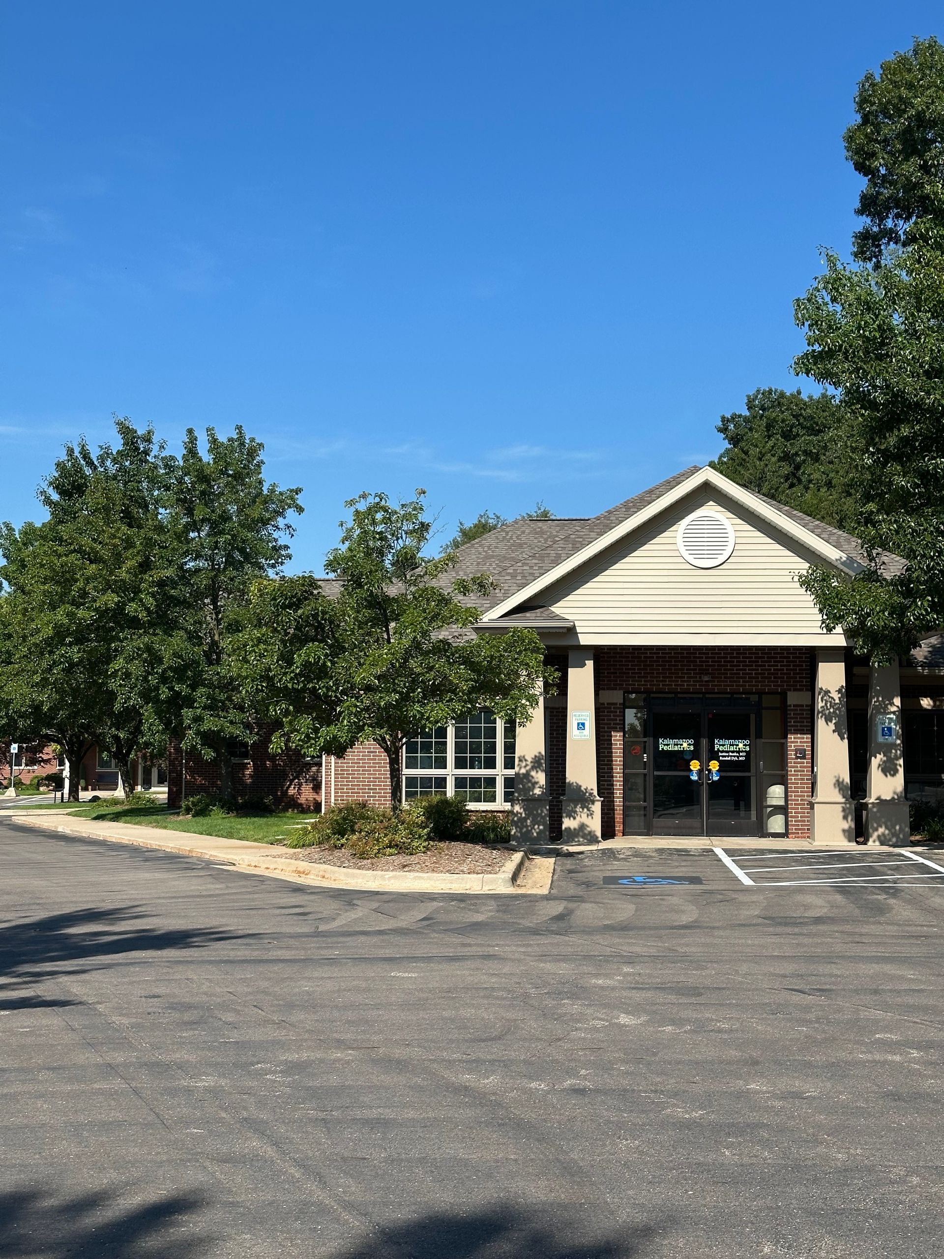 A low, beige building with a gabled roof, trees, and a paved driveway under a blue sky.