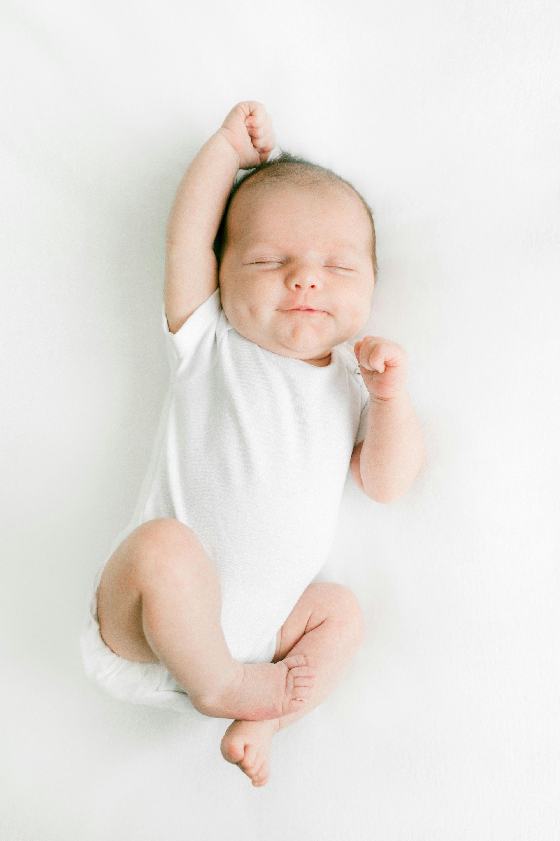 Smiling newborn baby in white onesie, arms raised on white background.