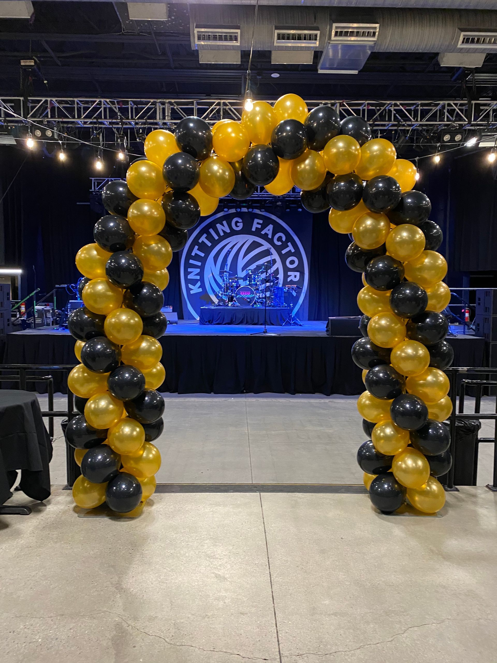 A large arch made of black and gold balloons in front of a stage.