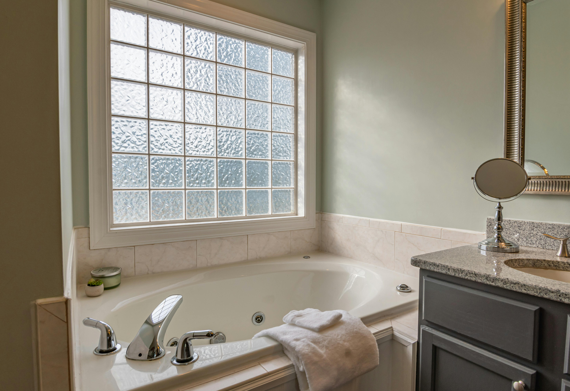 Bathroom with a bathtub, window, countertop, and mirror. Soft green walls, light-colored tile and a towel on the tub.