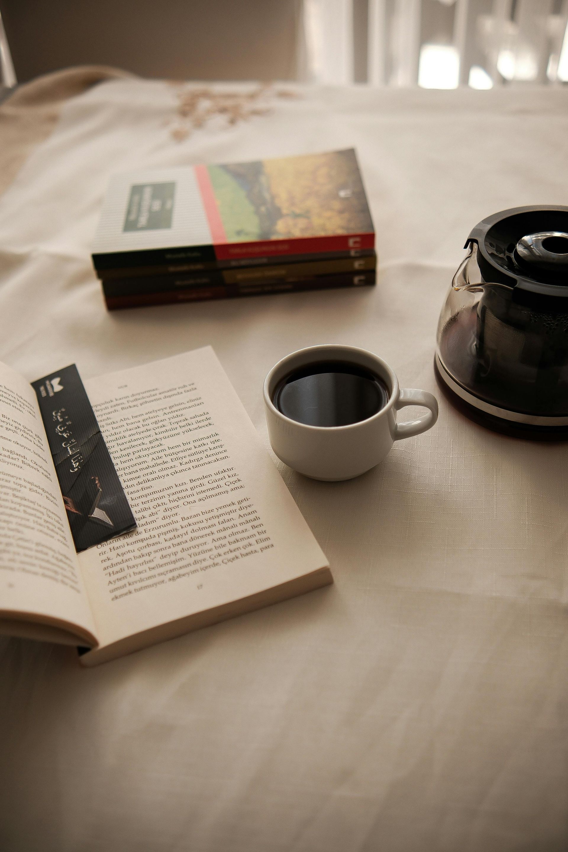 Open book with a bookmark, coffee cup, books, and coffee pot on a white tablecloth.