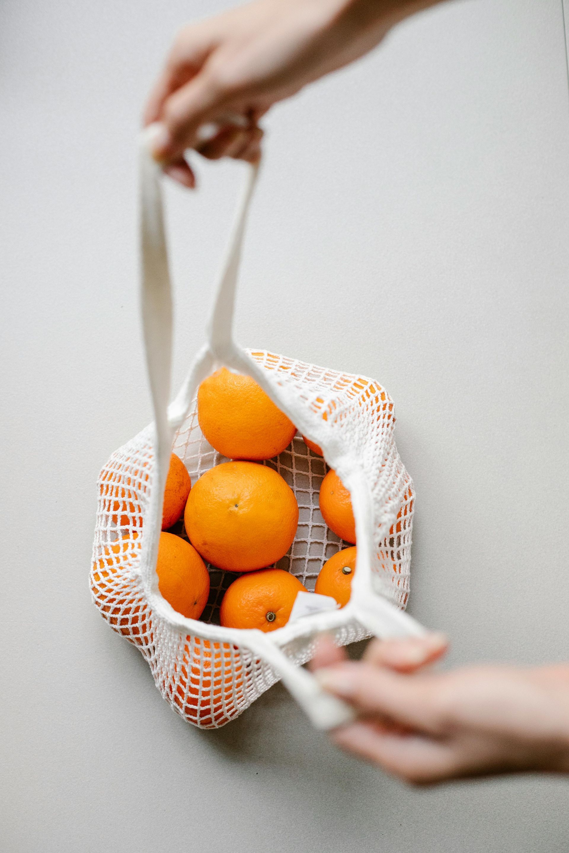 Hands holding a reusable white mesh bag filled with oranges against a neutral gray background.