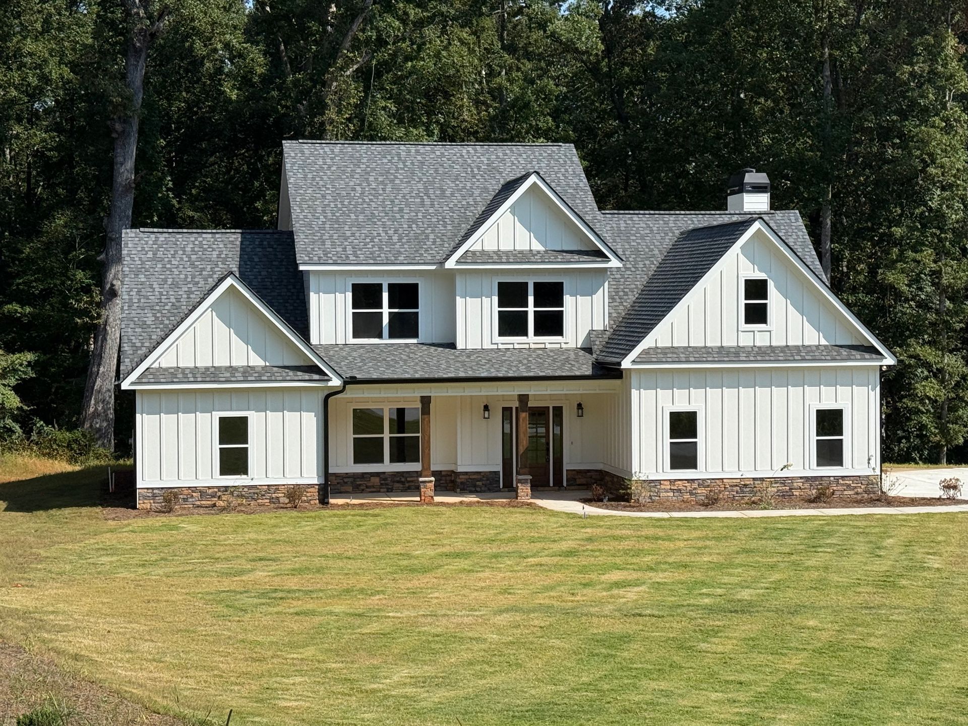 White two-story farmhouse with gray roof, set on a grassy lawn, surrounded by trees.