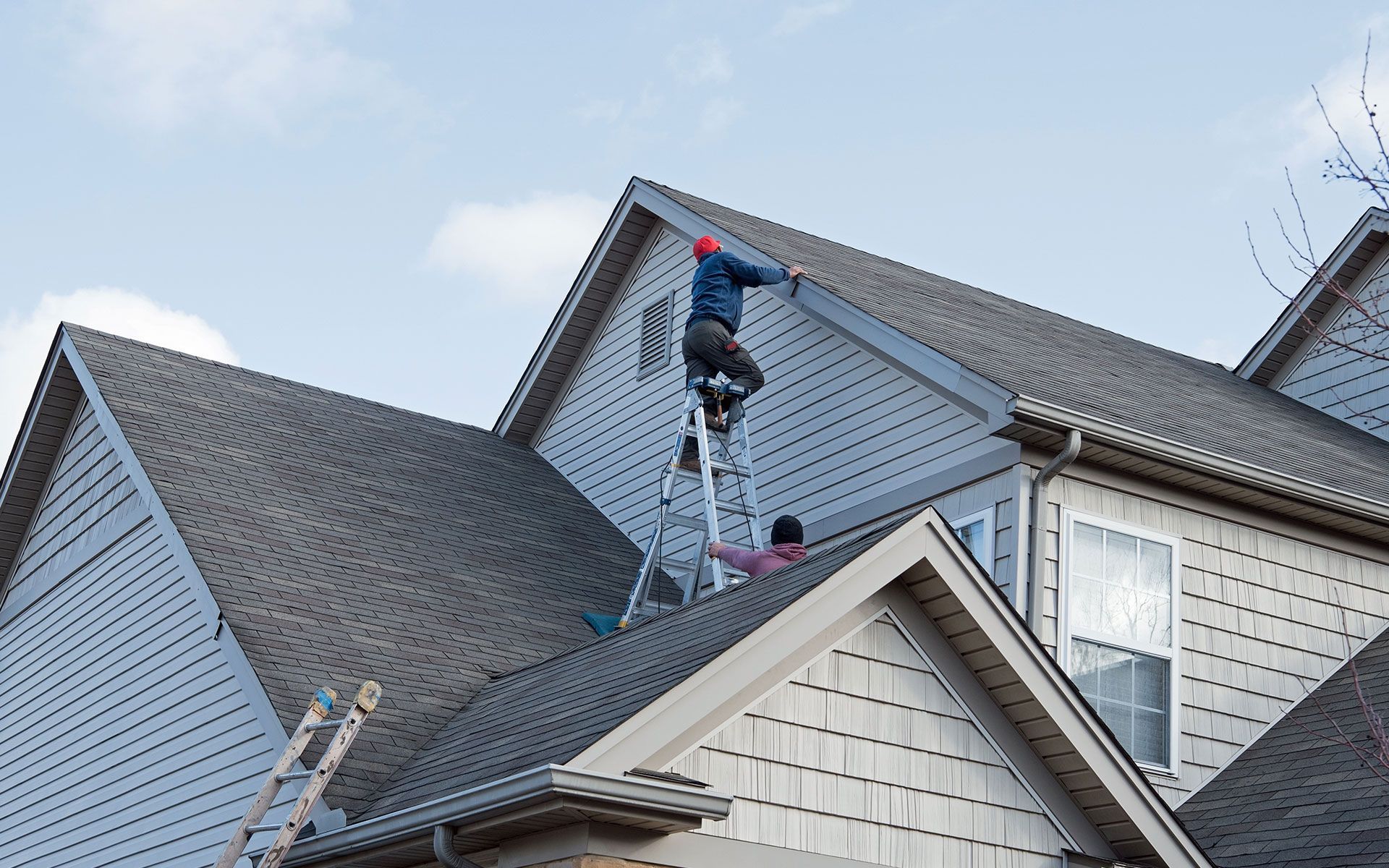 Two people on a roof; one on a ladder, and another on the roof installing siding. Blue sky in background.