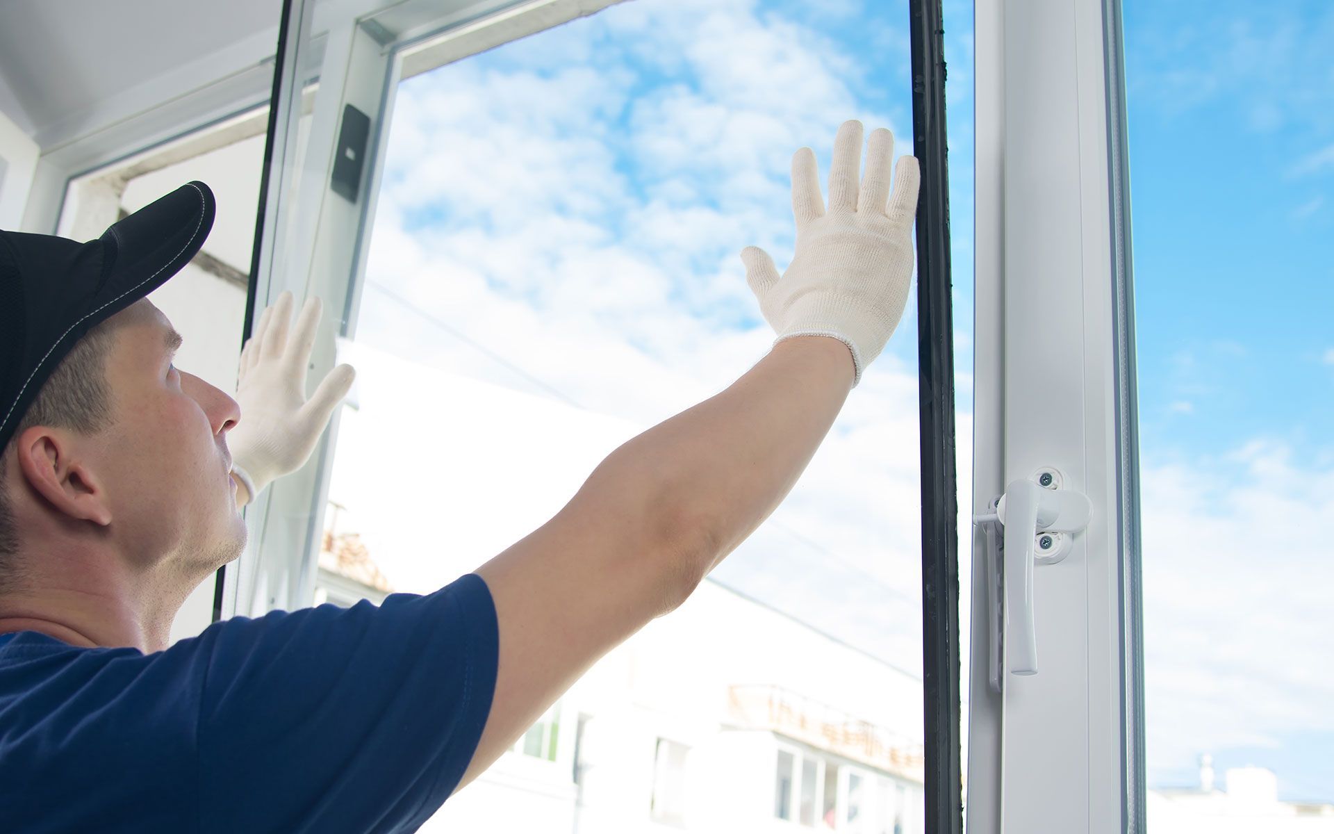 Man in a cap and gloves installing a window, arms outstretched, against a blue sky.