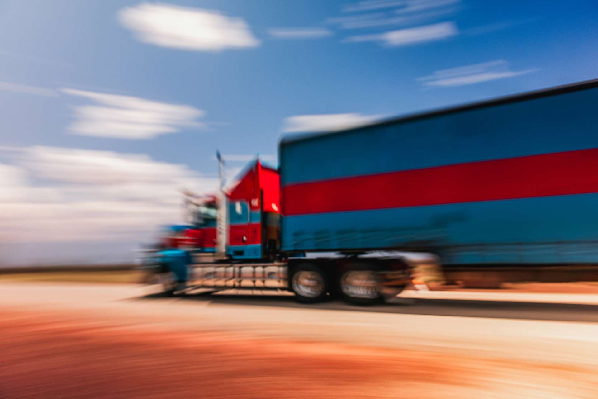 Blurred semi-truck with blue and red trailer driving under a partly cloudy sky. Blurred semi-truck with blue and red trailer driving under a partly cloudy sky.