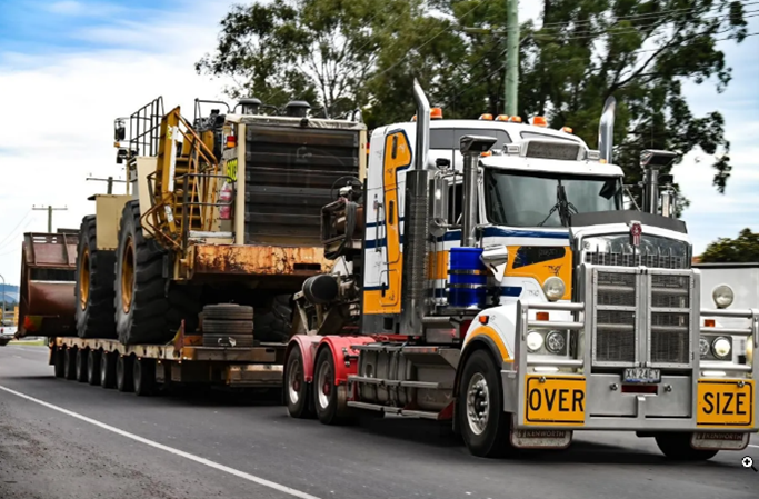 An over-size truck with a large trailer traveling down a road surrounded by greenery.