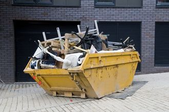 A yellow dumpster filled with trash is parked in front of a brick building.