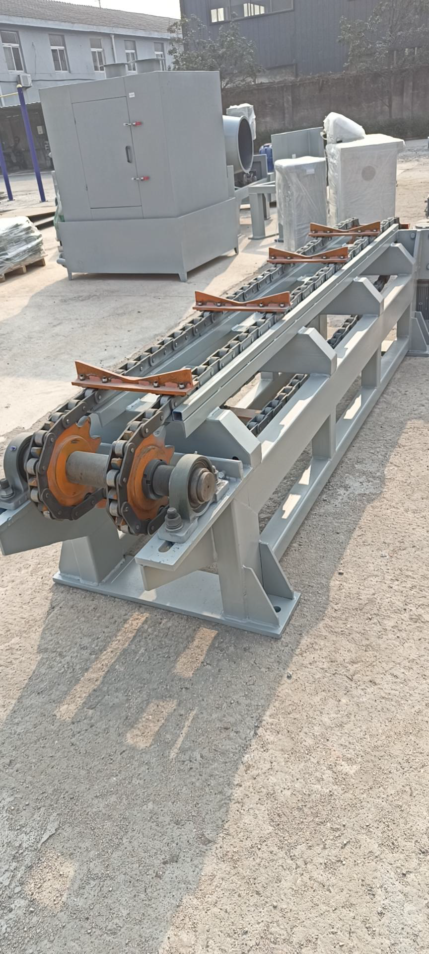 Industrial machinery with chains and gears sits on gravel. Buildings in the background.