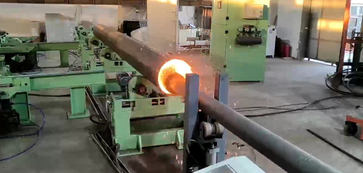 A long metal pipe is heated in a factory setting, with bright orange glowing inside the pipe and sparks flying.