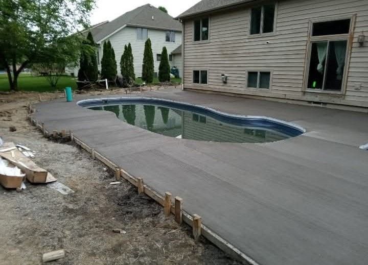 Two wooden lounge chairs with white cushions beside a blue swimming pool in a garden setting.