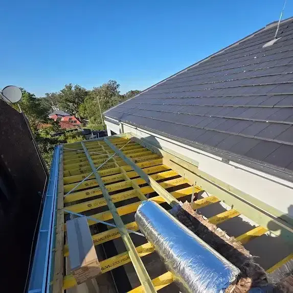 Rooftop under construction: wooden beams, insulation, and rolled silver foil. Another roof nearby, blue sky — McLuckie Metal Roofing In Lugarno, NSW