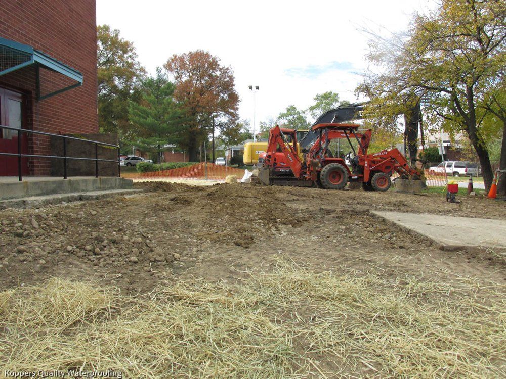 A red tractor is working on a dirt field in front of a brick building