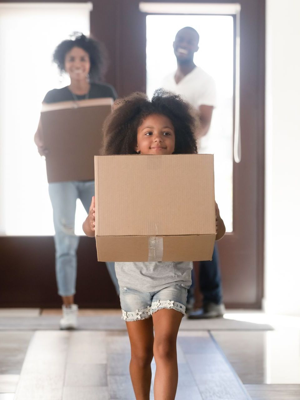 Family carrying boxes into a new home. Girl leads, smiling. Parents follow, doorway visible.
