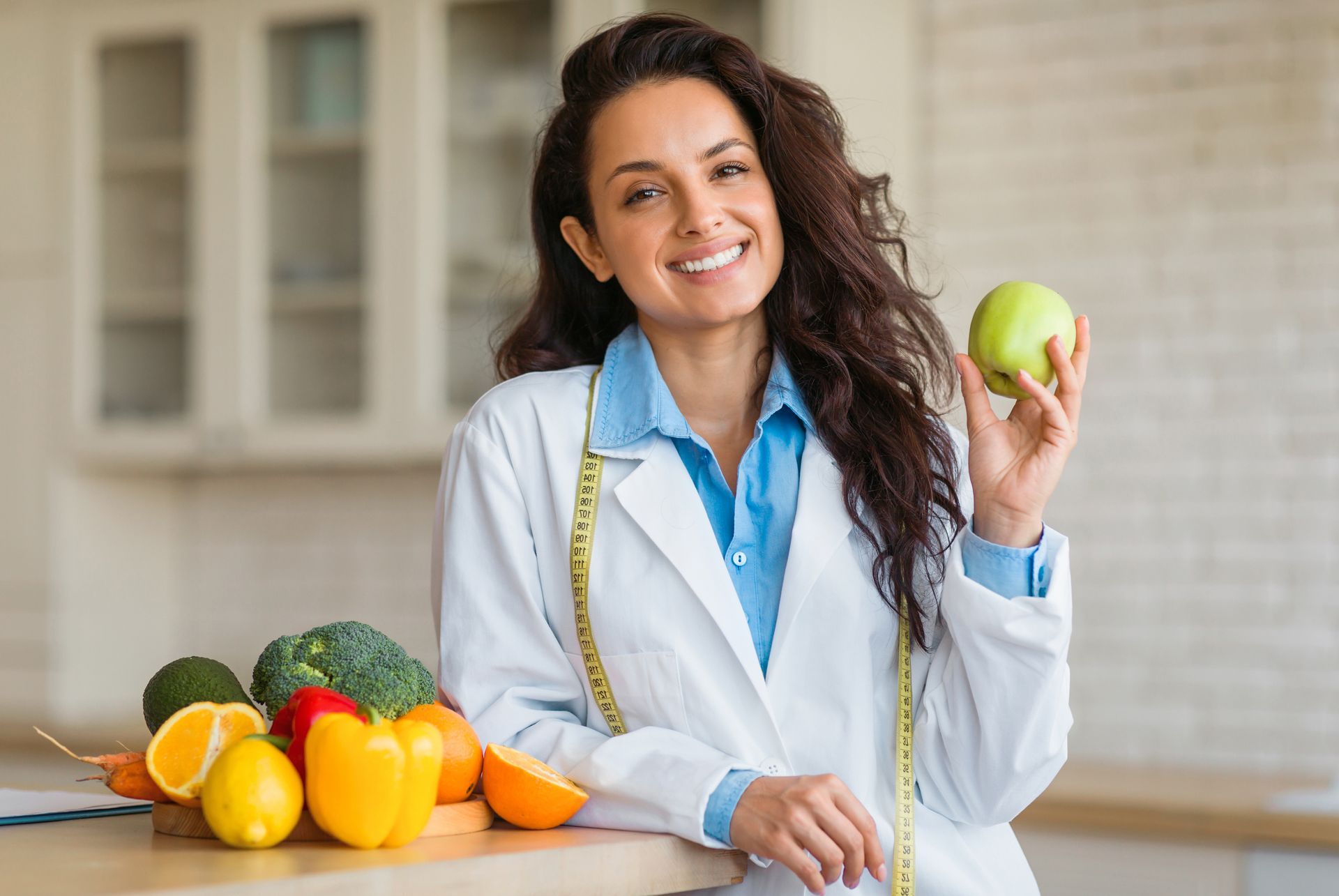 Dietitian in a white coat holds a green apple, smiling next to a table of fresh produce.