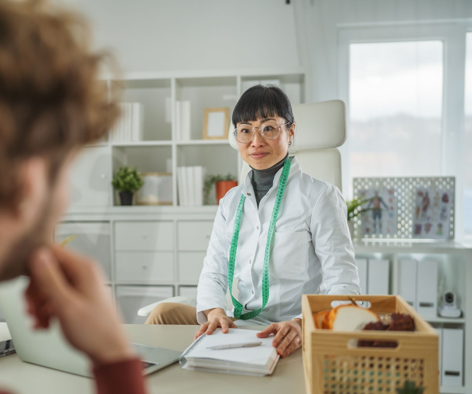 A dietitian in a white coat sits at a desk, looking at a patient. A measuring tape is around her neck.