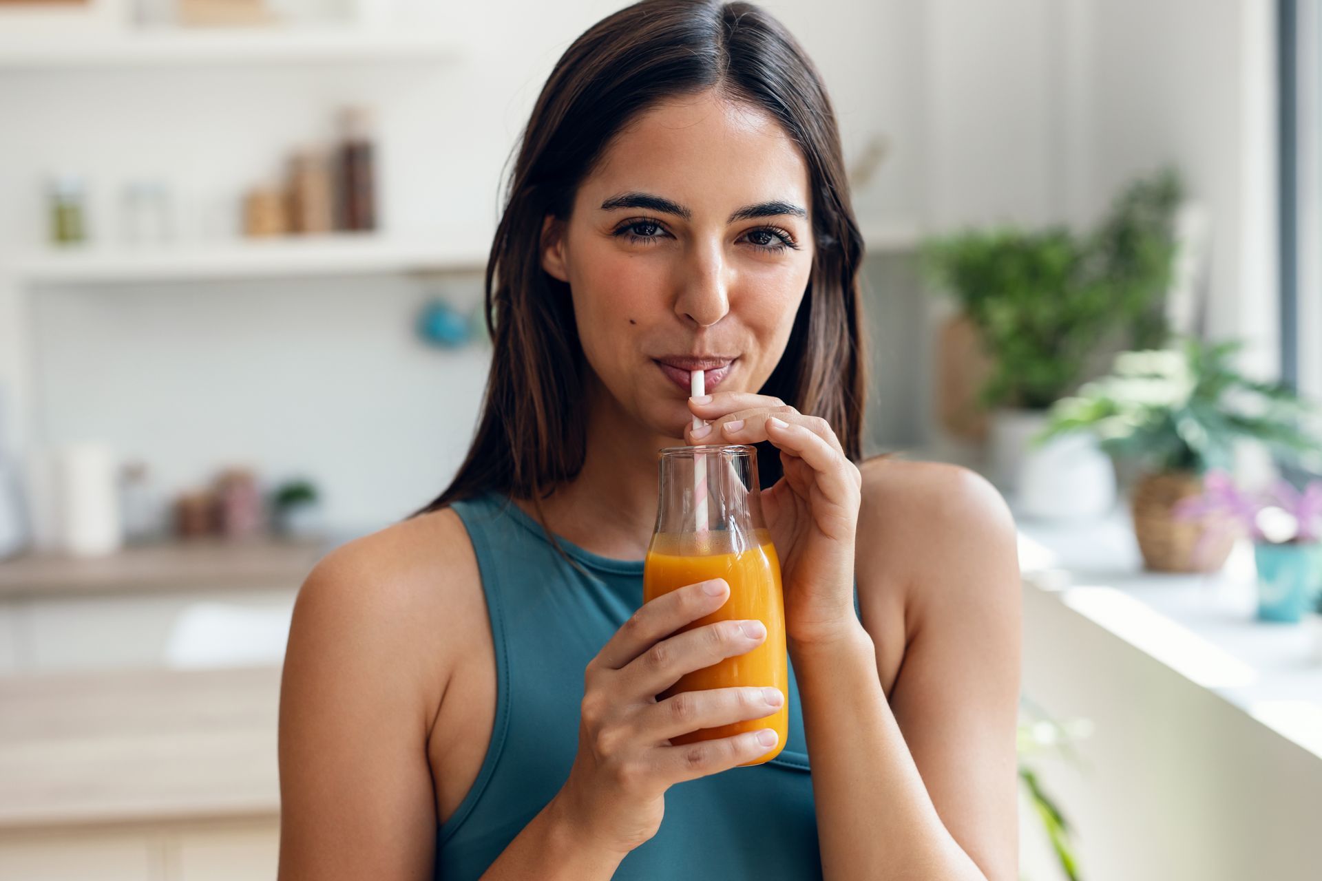 Woman drinks from orange juice bottle with straw in kitchen.
