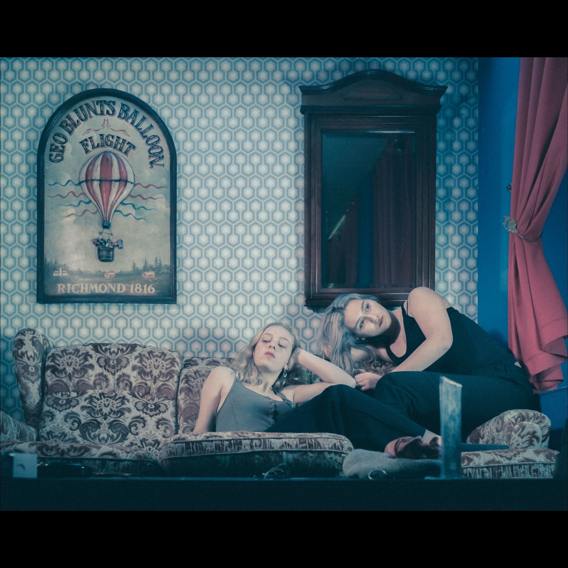 Two people resting on a patterned couch indoors, near a poster and a mirror.