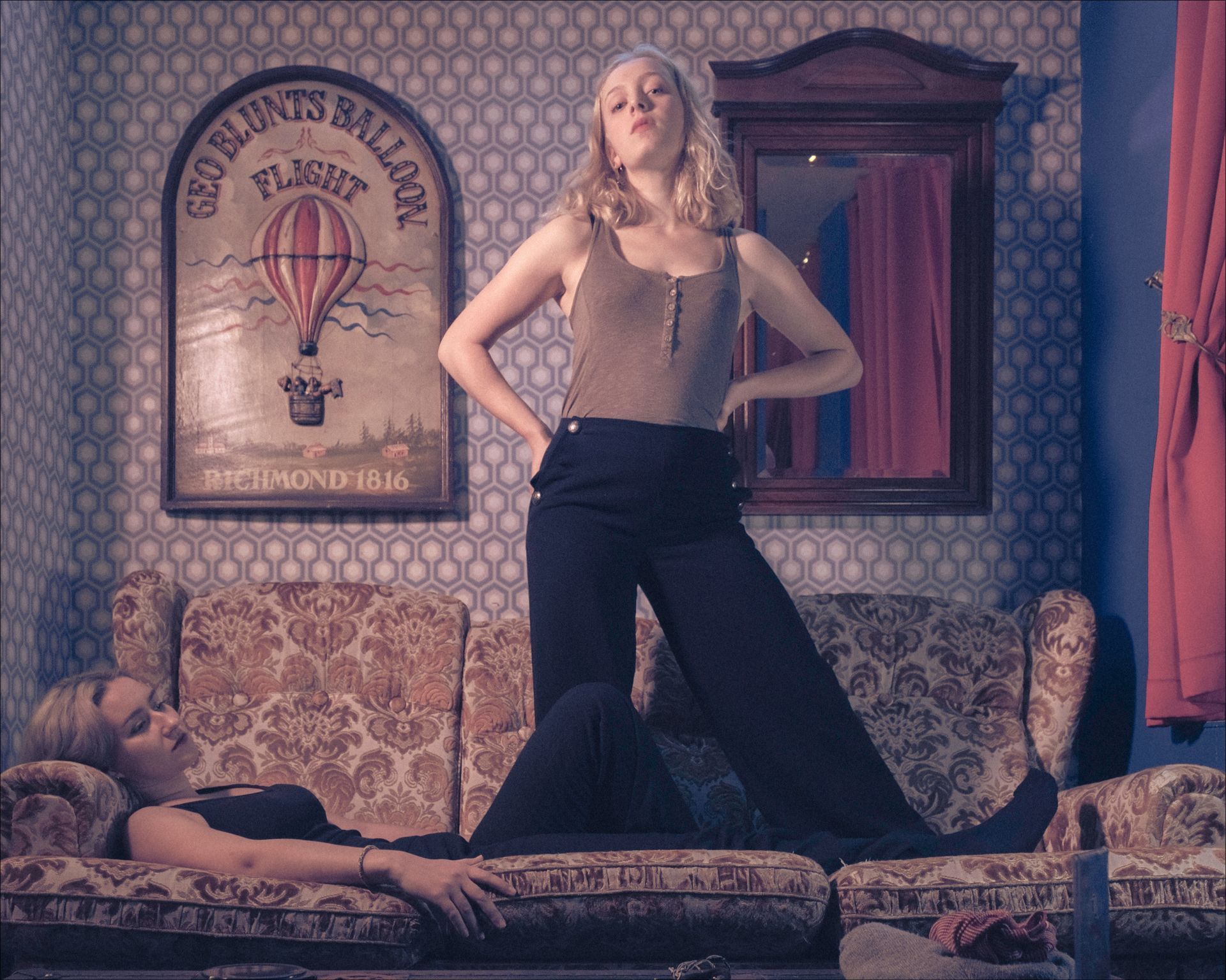 Woman standing over a woman on a couch. Vintage wallpaper, a hot air balloon poster, and a mirror are in the background.