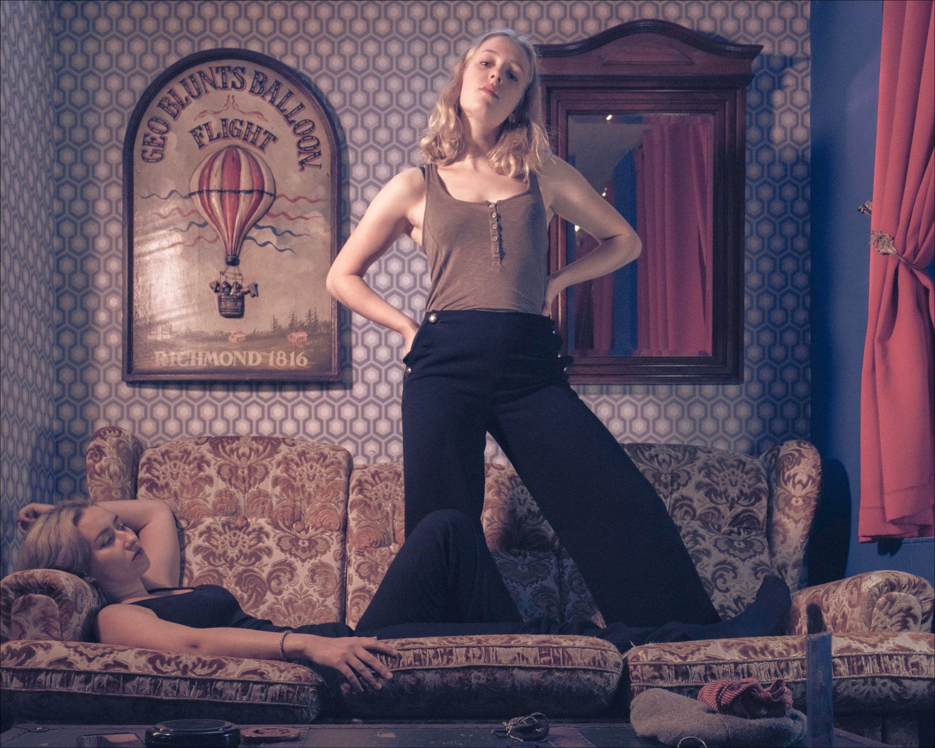 Woman standing on couch, posing. Another woman reclines on the couch. Interior, hot air balloon art.