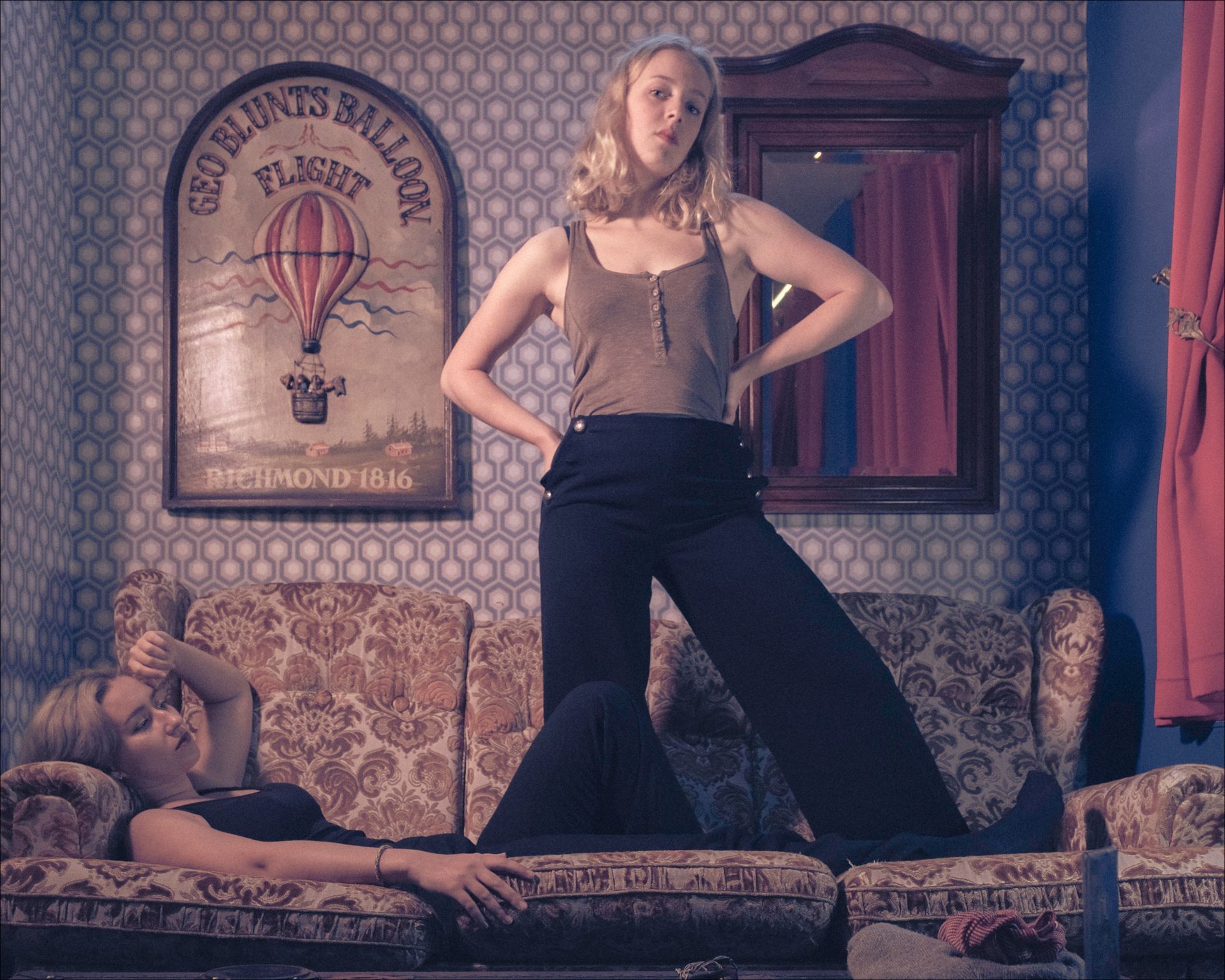 Two women in vintage room; one standing, one reclining on a floral couch. Hot air balloon poster on wall.