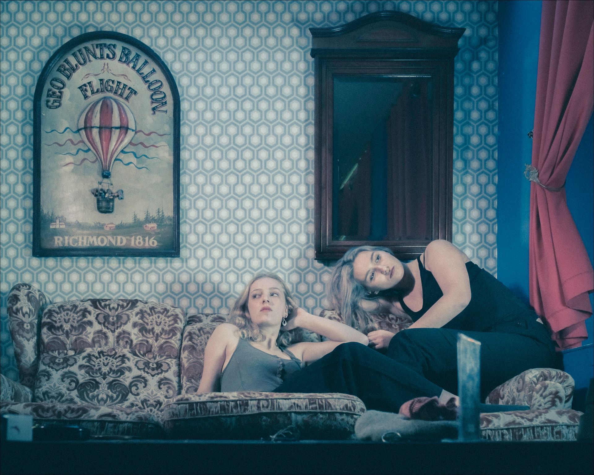 Two women on a patterned sofa, one gazing up, the other leaning, against a wall with a mirror and print.