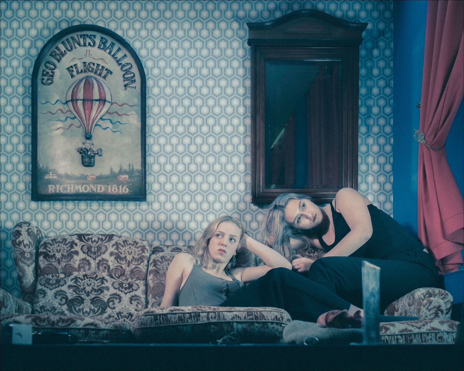 Two people relax on a floral couch, looking at a hot air balloon poster in a room with patterned wallpaper.