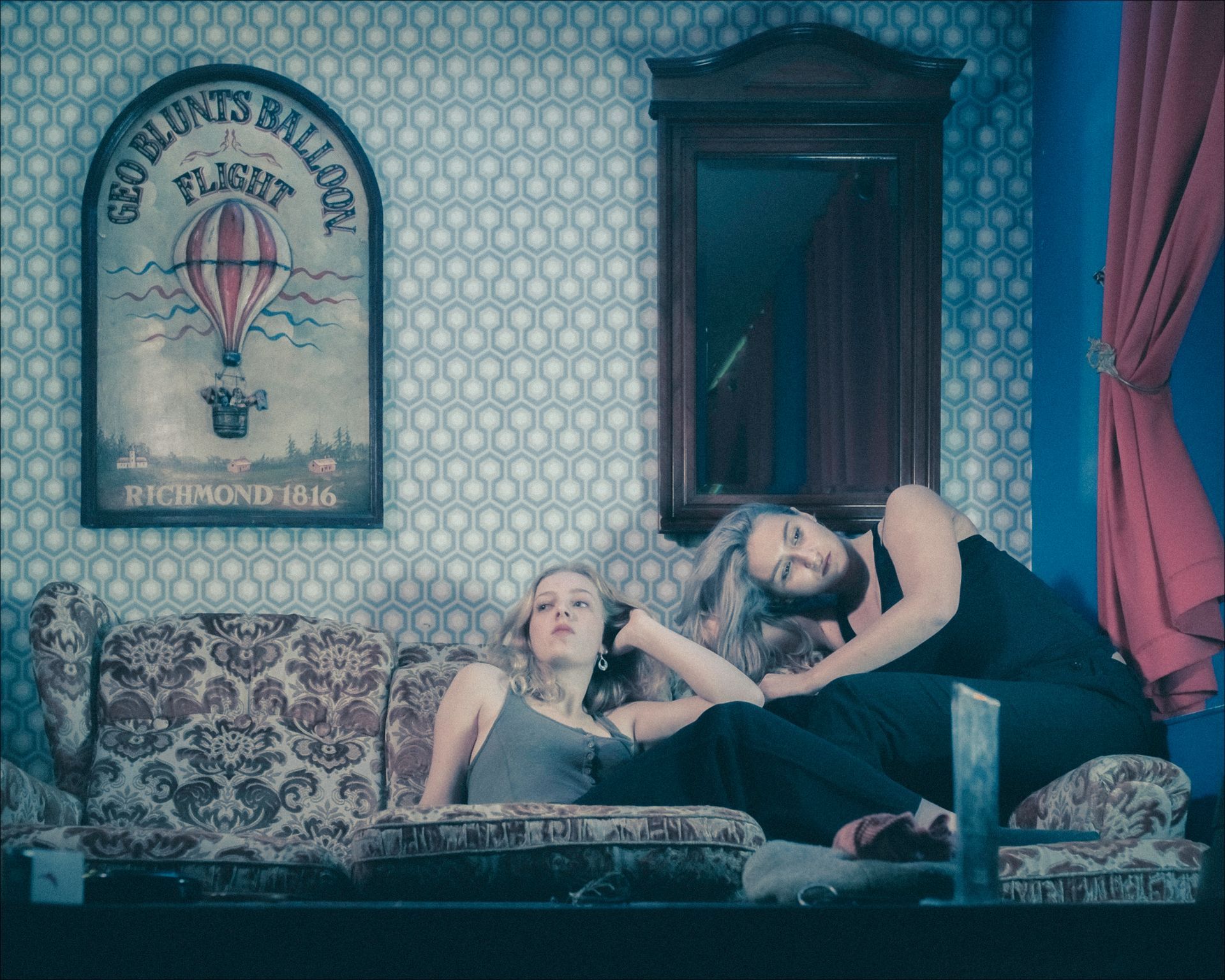 Two people recline on a patterned couch, near a poster and a mirror. One person rests their head on the other's shoulder.